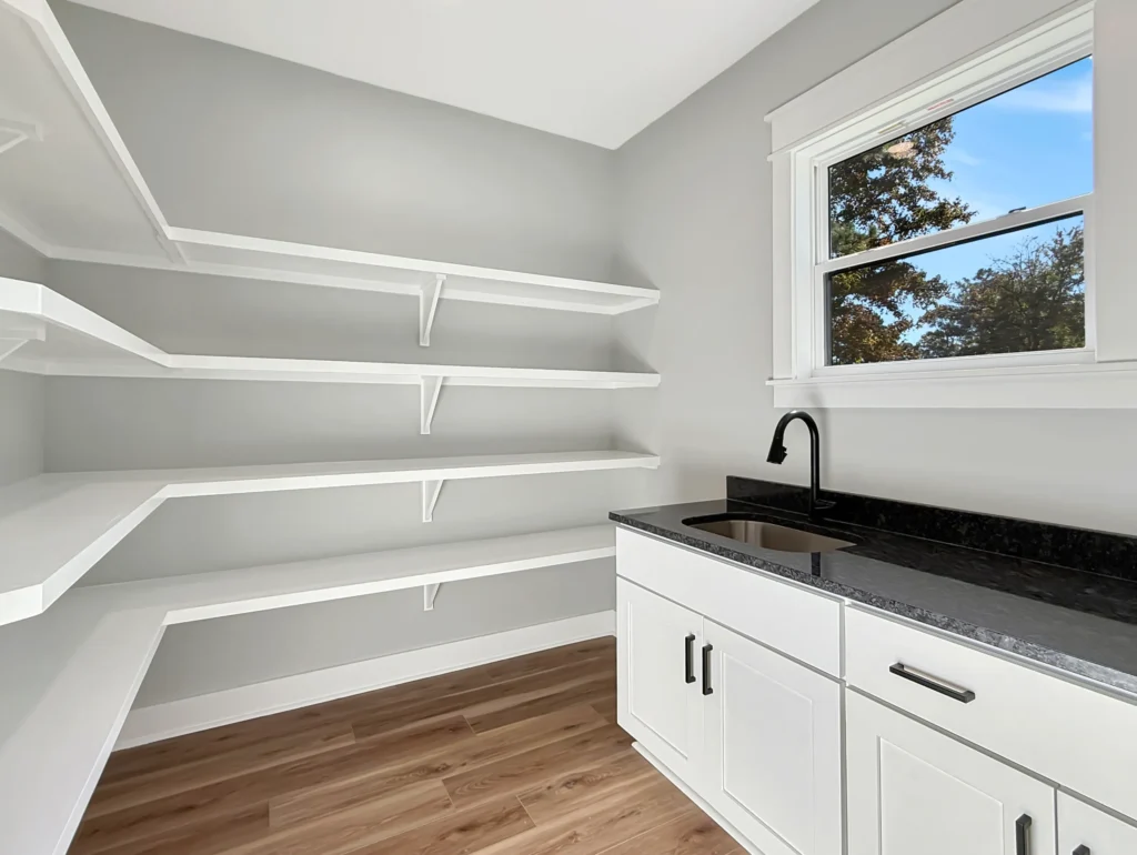 A spacious walk-in pantry with wrap-around white shelving and a utility sink with black granite countertops in an Elizabeth City new construction home.